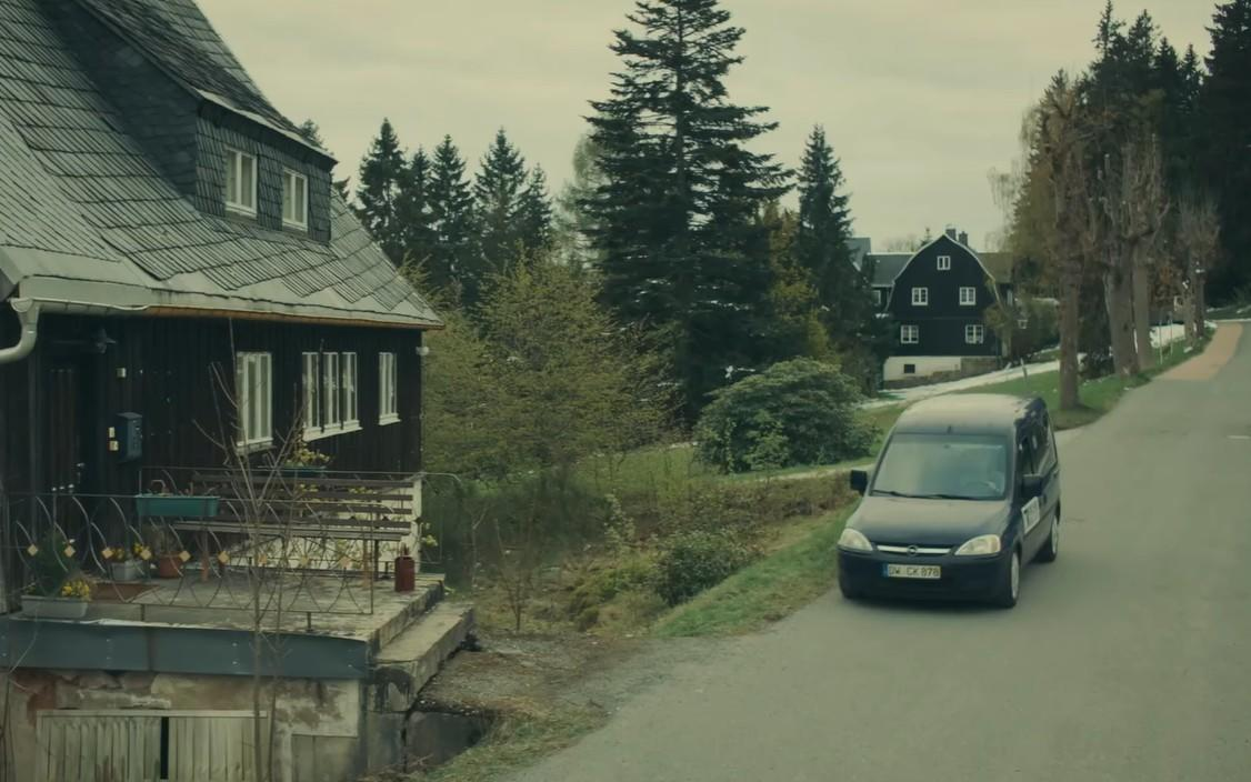 Dunkelblaues Auto fährt auf einer Straße an einem Haus mit Wald im Hintergrund vorbei.