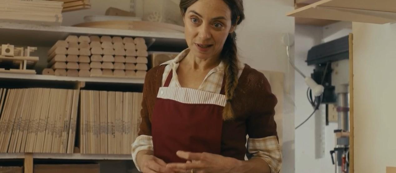 Eine Frau mit einem roten Latz und kariertem Hemd steht vor Regalen mit Holz.