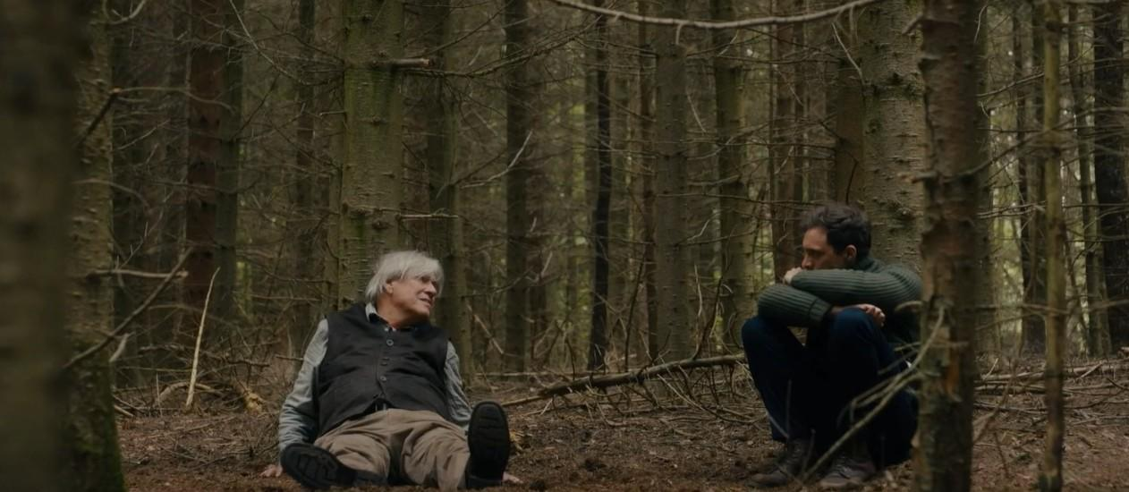 Zwei Männer sitzen auf dem Waldboden, umgeben von Bäumen. Einer sitzt mit ausgestreckten Beinen, der andere kauert.