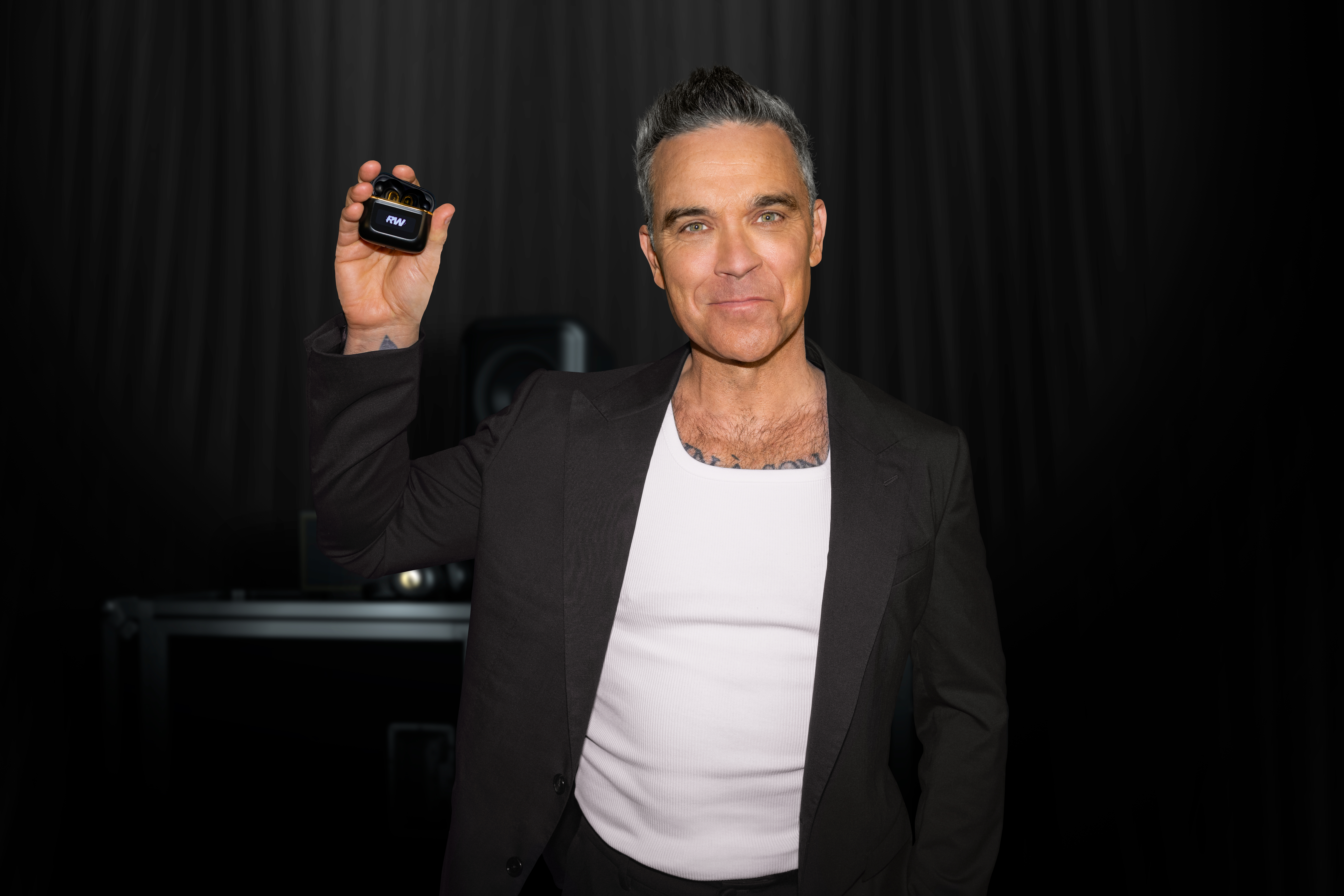 Robbie Williams egy fekete töltőtokot tart fejhallgatókkal és RW logóval.