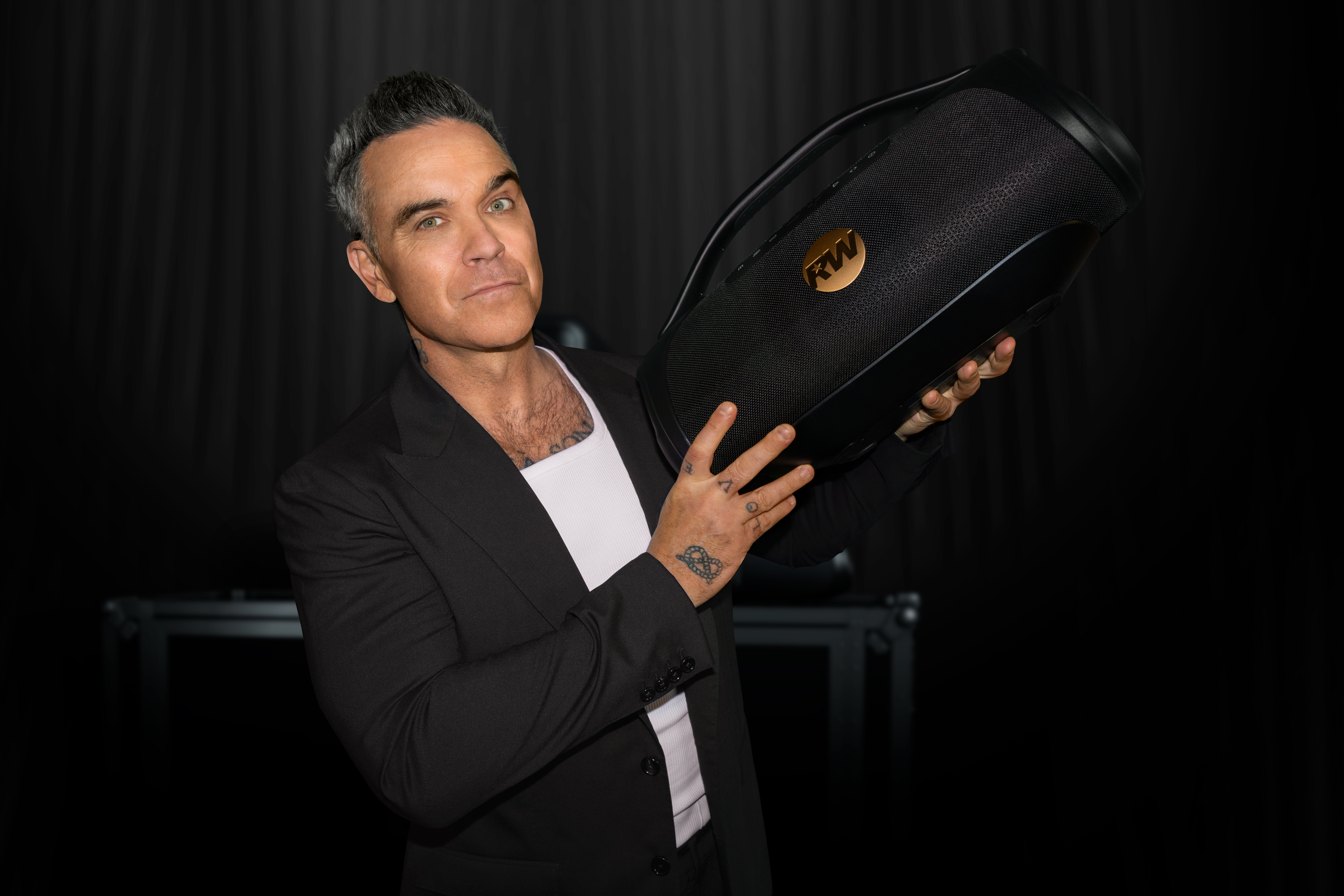 Robbie Williams fekete öltönyben, fekete hangszórót tart RW logóval.