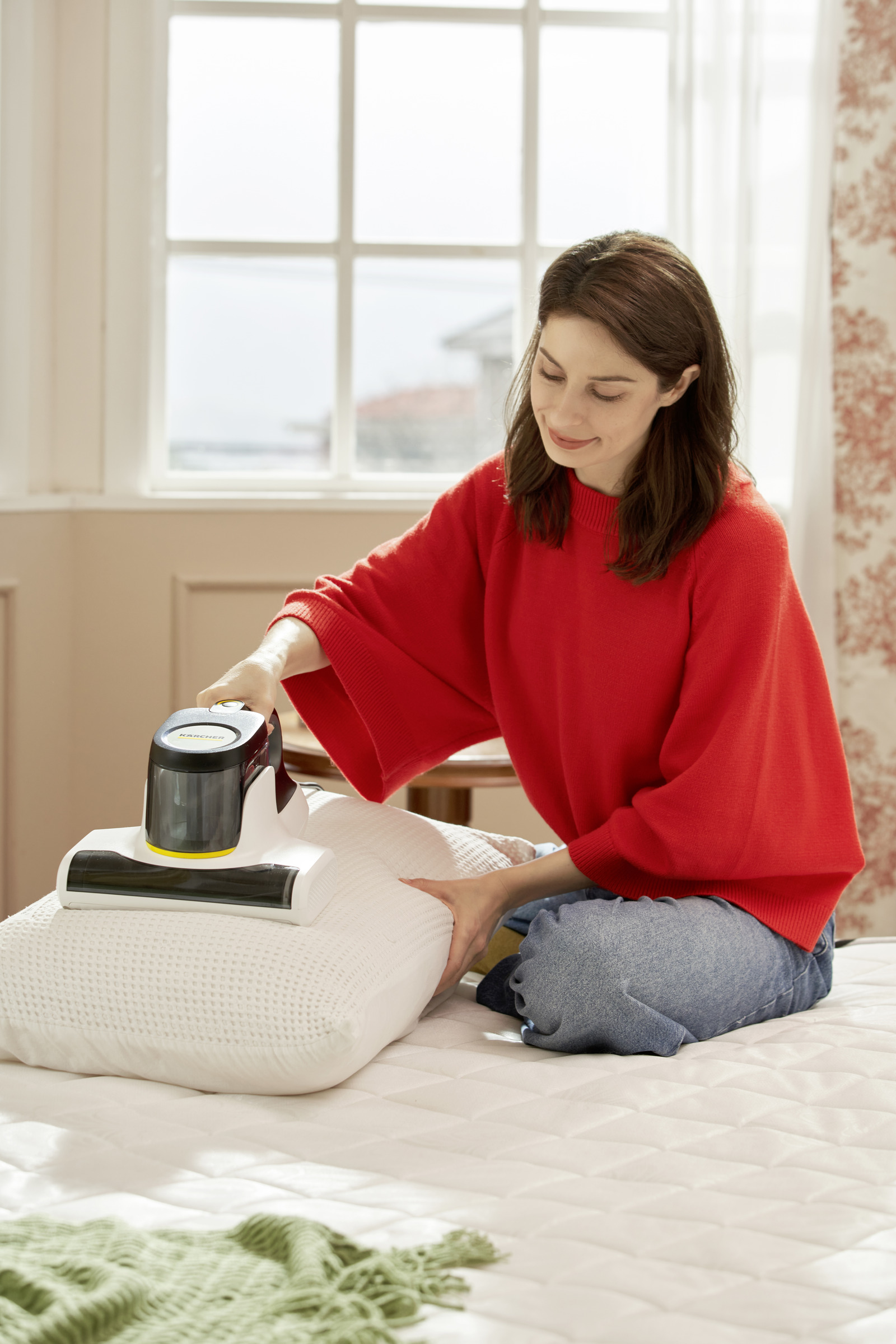 Kobieta w czerwonym swetrze odkurza białą poduszkę na łóżku.