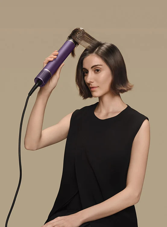 Kobieta stylizuje brązowe włosy fioletową prostownicą na beżowym tle.