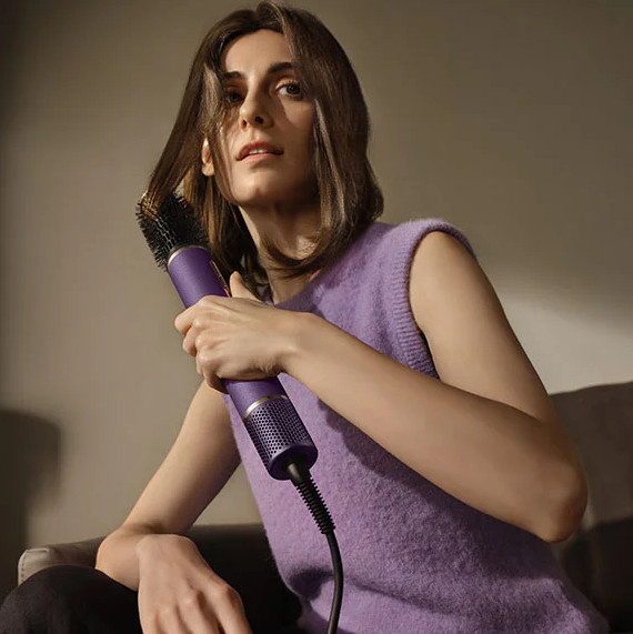 Kobieta z brązowymi włosami stylizuje je fioletową suszarką z okrągłą szczotką.