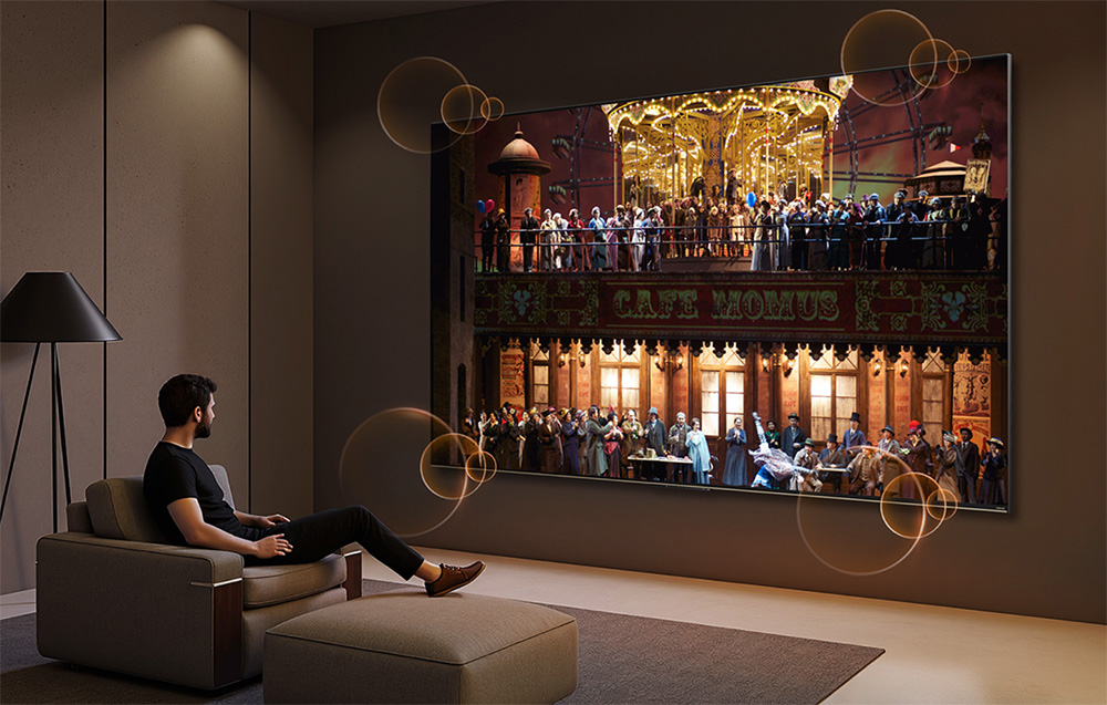 Mężczyzna siedzi na sofie, ogląda duży ekran z sceną operową i karuzelą.