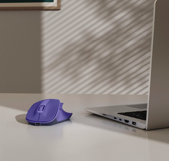 Fioletowa mysz komputerowa obok otwartego laptopa na białym biurku.