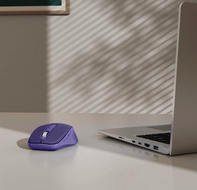 Fioletowa mysz obok laptopa na białym biurku, z cieniami na ścianie.