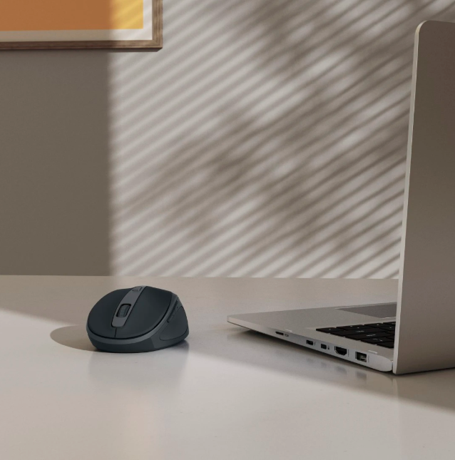 Ciemnoszara mysz obok otwartego laptopa na białym biurku.