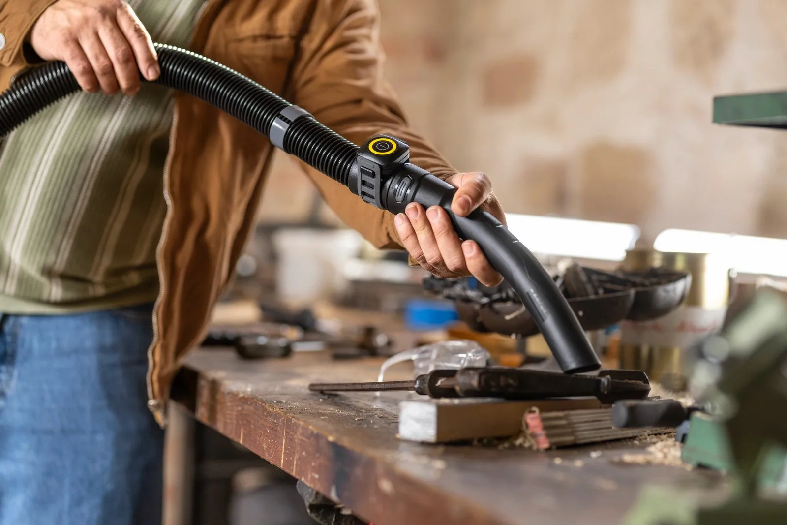 Osoba czyści stół warsztatowy czarną rurą odkurzacza, narzędzia porozrzucane.