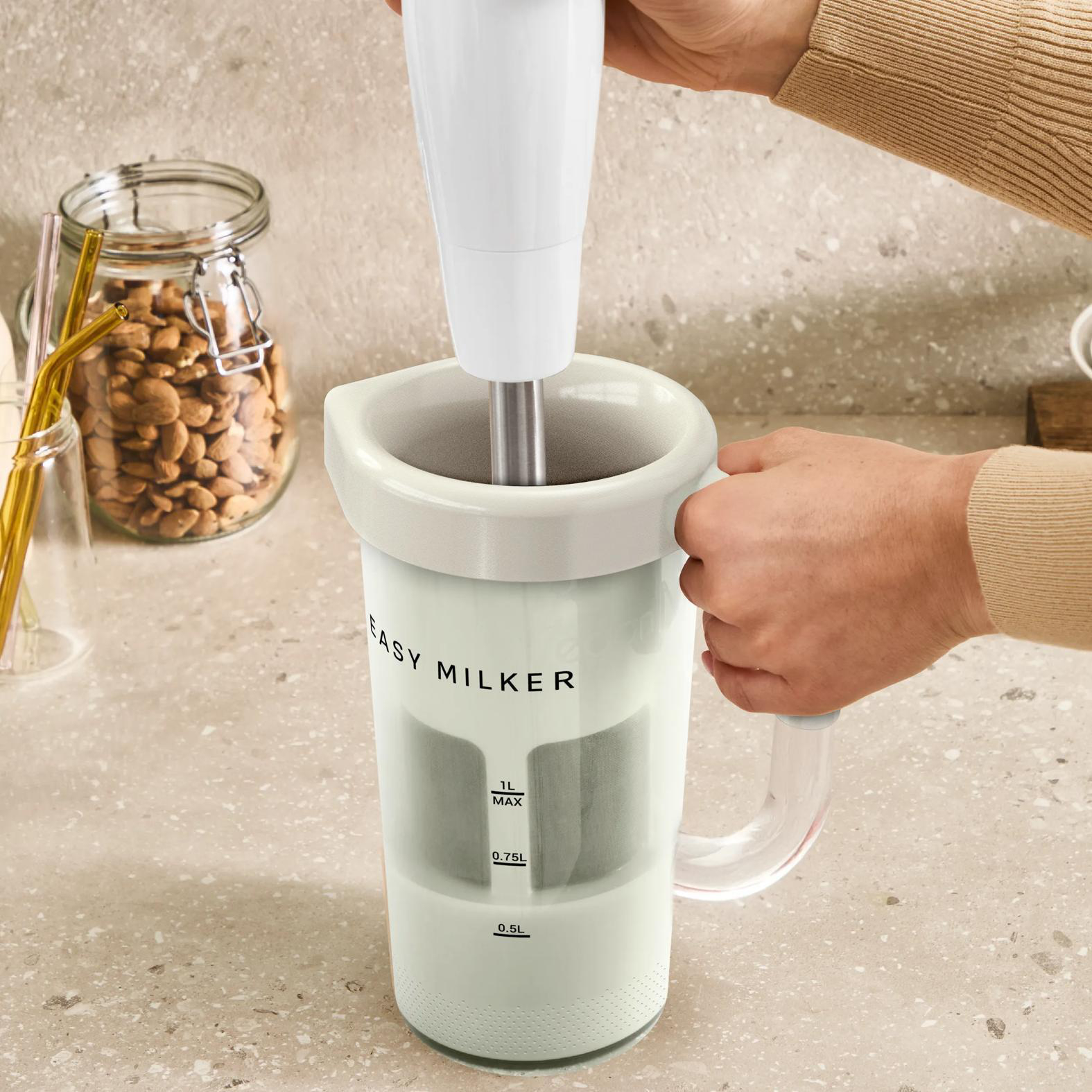 Eine weiße Milchmaschine mit einem Stabmixer wird von Händen gehalten. Daneben ein Glas mit Nüssen.