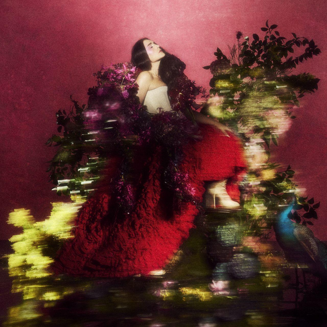 Frau in rotem Kleid sitzt inmitten von Blumen, Pfau rechts.