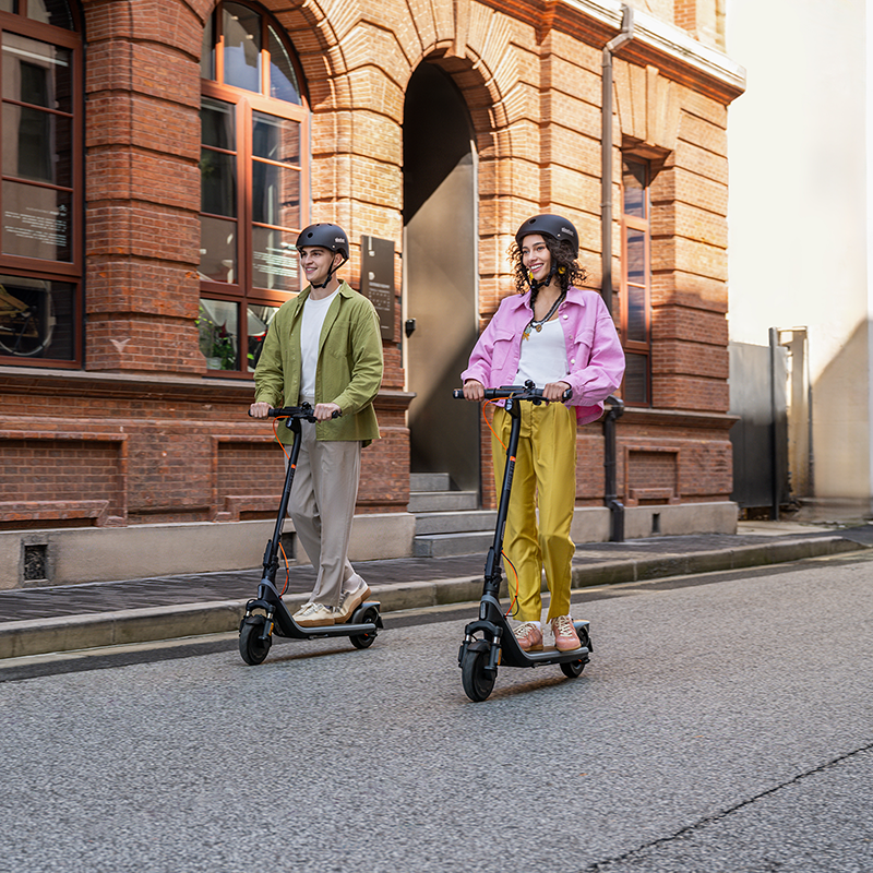 İki kişi tuğla binanın önündeki sokakta elektrikli scooter sürüyor.