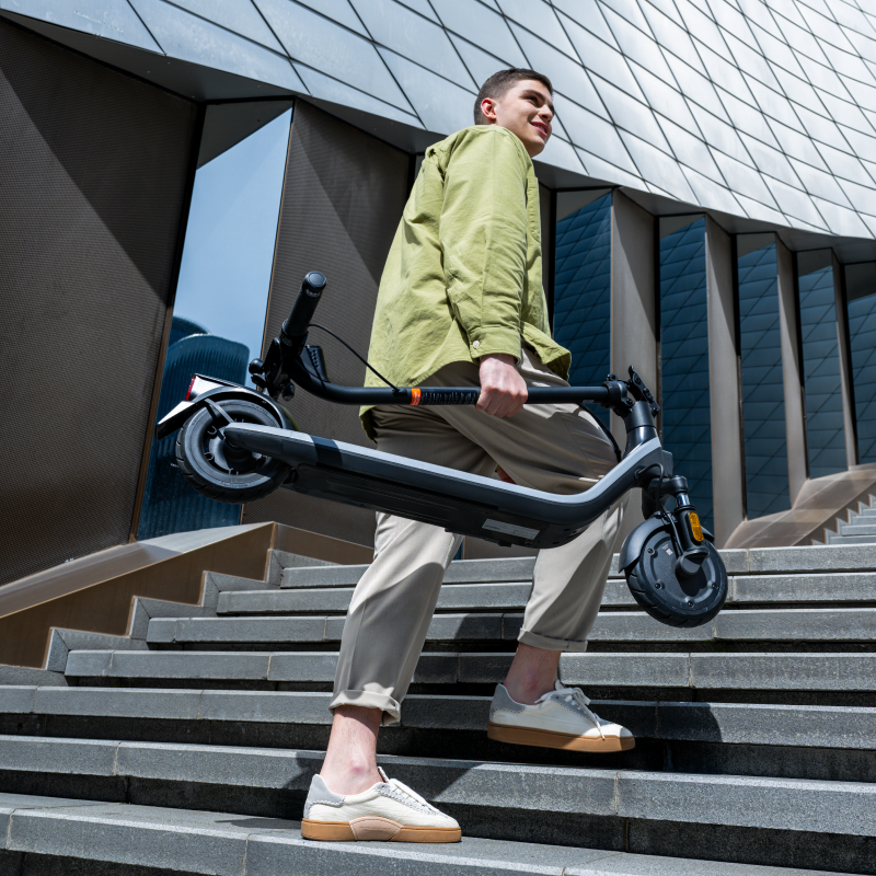 Genç adam katlanmış elektrikli scooterı beton merdivenlerden yukarı taşıyor.