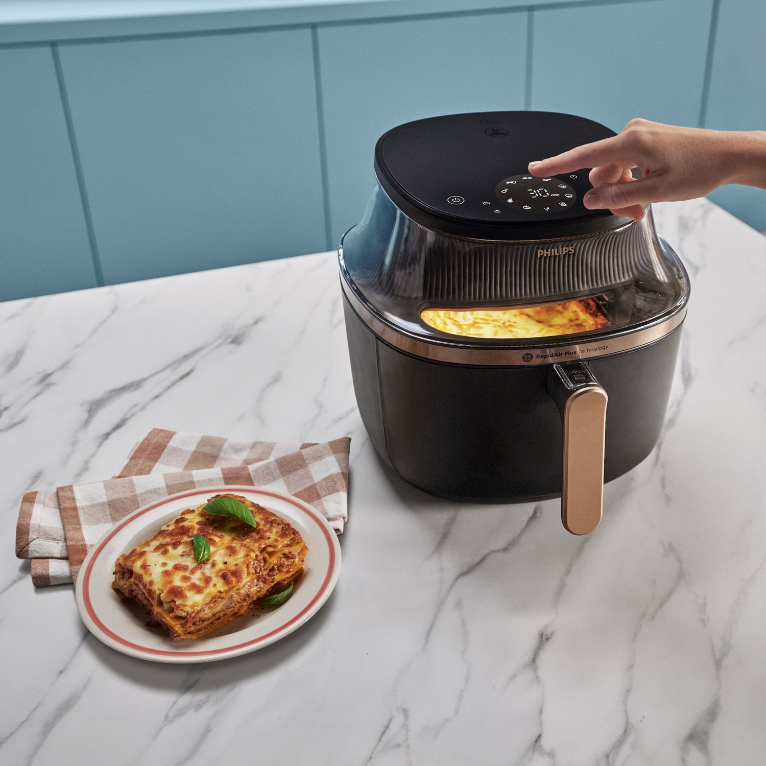 Una mano regola una friggitrice ad aria accanto a una fetta di lasagne su un piatto.