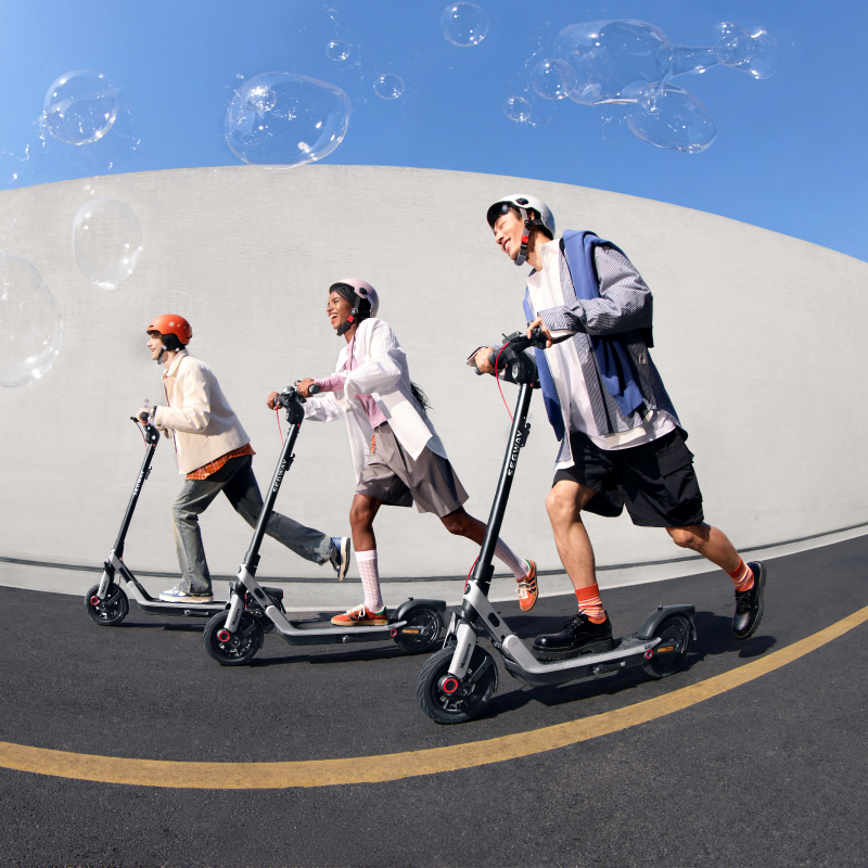 Elektrikli scooter kullanan üç kişi gökyüzünde balonlarla bir sokakta sürüyor.