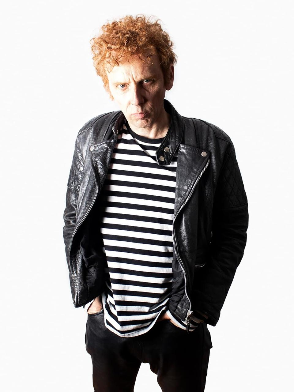 Mann mit roten Locken, Lederjacke, gestreiftem Shirt und Hosen.