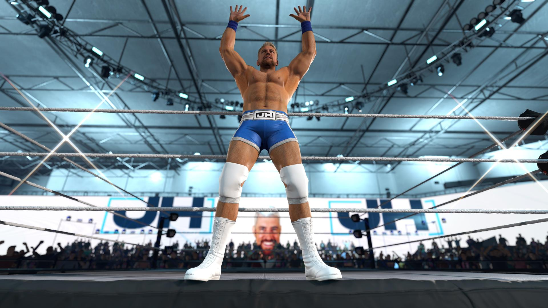 Ein Wrestler in blauer Hose mit JH-Logo steht im Ring mit erhobenen Armen.
