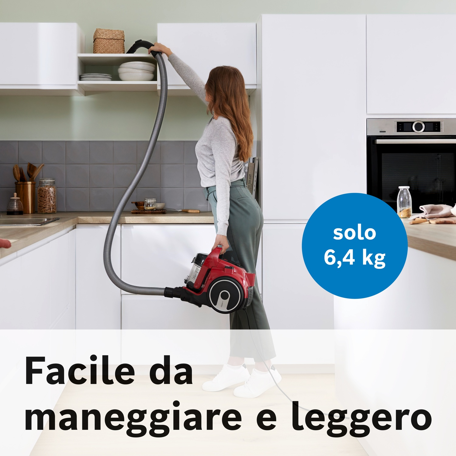 Donna in maglione grigio e pantaloni verdi aspira cucina con aspirapolvere rosso.