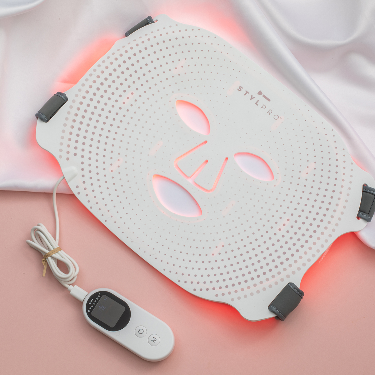 Una mascarilla facial LED blanca con luces rojas descansa sobre satén y fondo rosa.