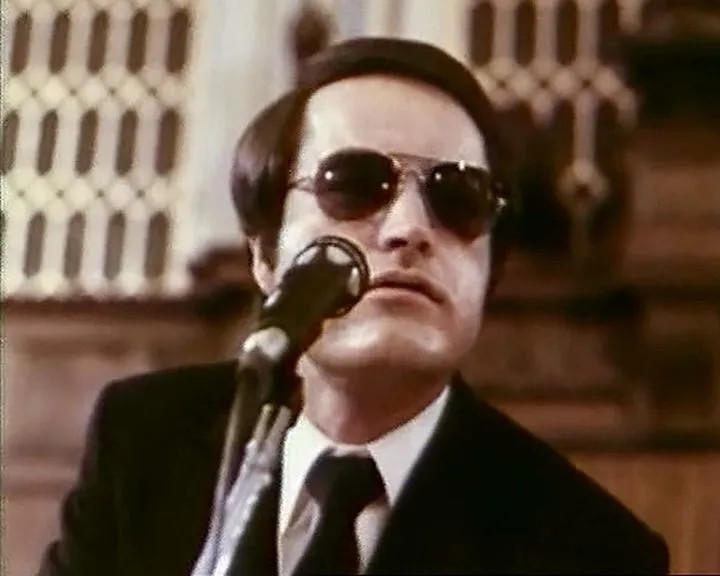 Mann in Sonnenbrille spricht ins Mikrofon, Anzug und Krawatte.