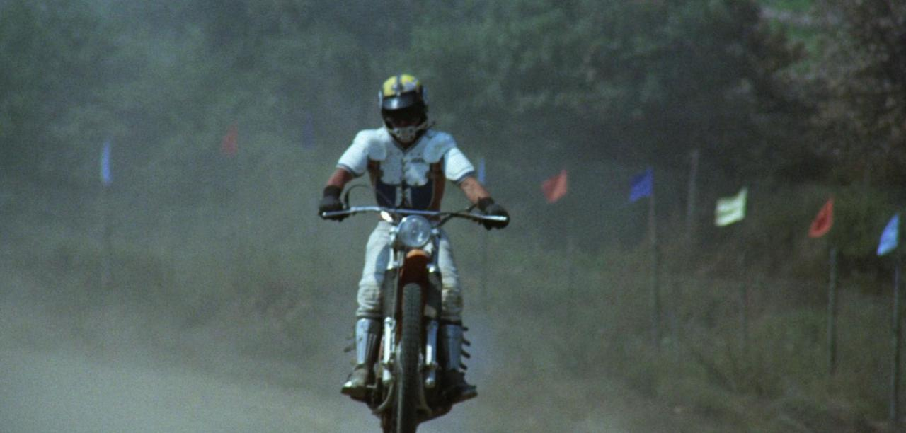 Ein Motorradfahrer in voller Montur fährt auf einem staubigen Feld mit bunten Flaggen.