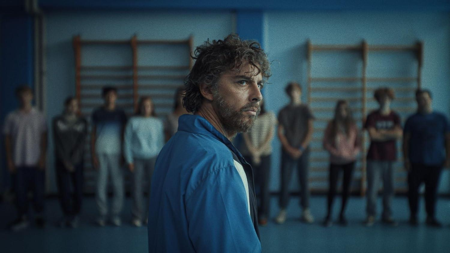 Ein Mann mit lockigem Haar in blauer Jacke blickt nach rechts, Schüler dahinter.