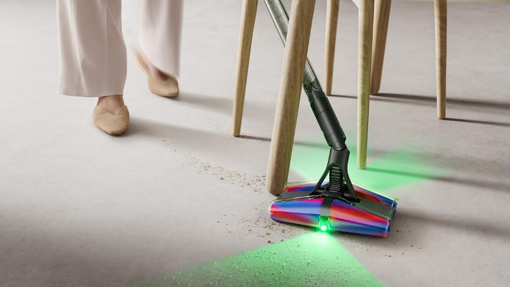 Odkurzacz z zielonymi światłami pod drewnianymi krzesłami, przechodzi osoba w beżowych spodniach i butach.