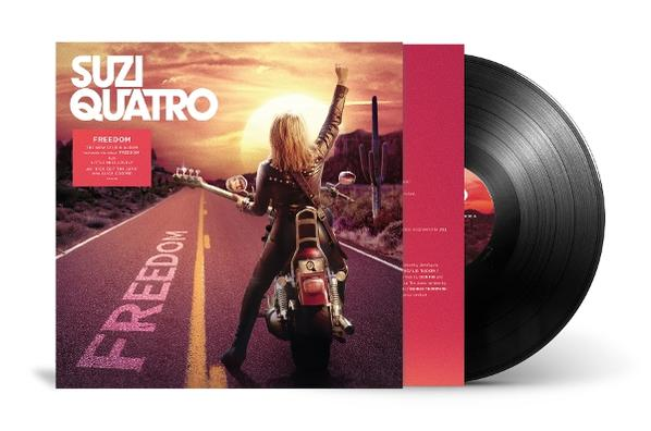 Suzi Quatro auf einem Motorrad auf einer Straße bei Sonnenuntergang, Gitarre haltend.