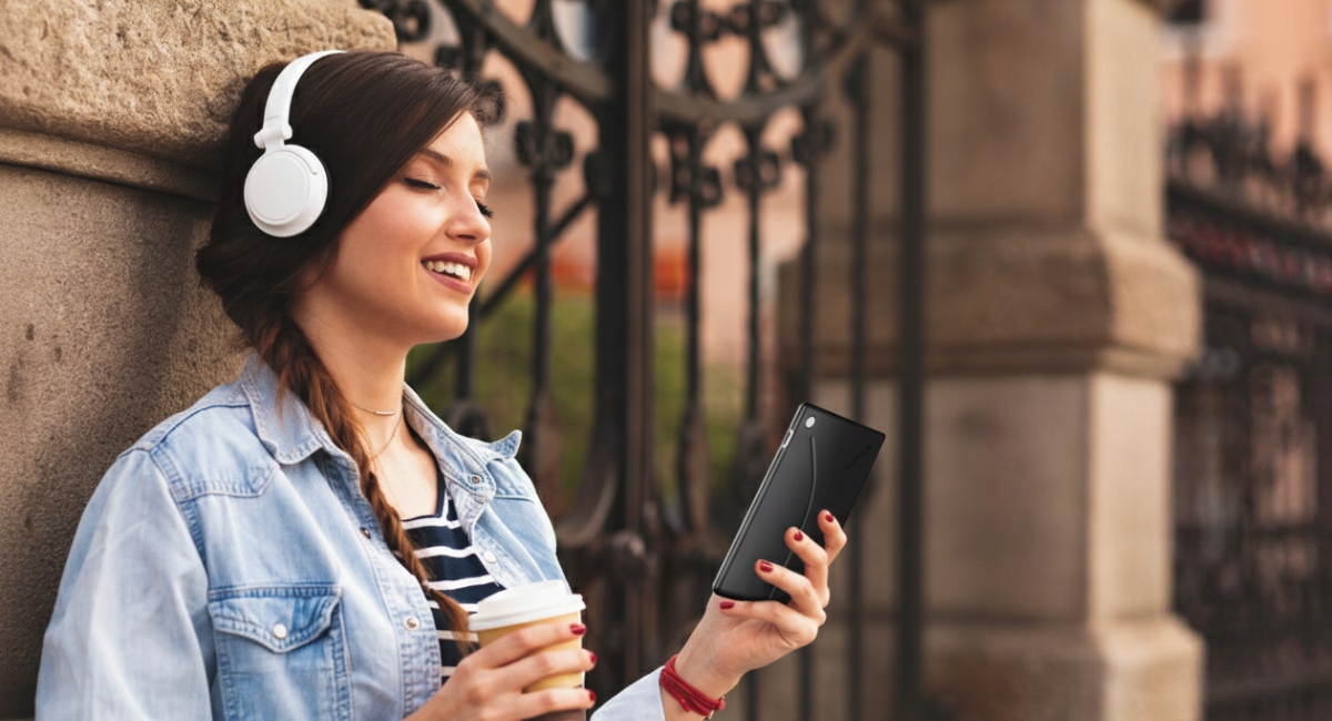 Młoda kobieta ze słuchawkami i kawą, przewija telefon.