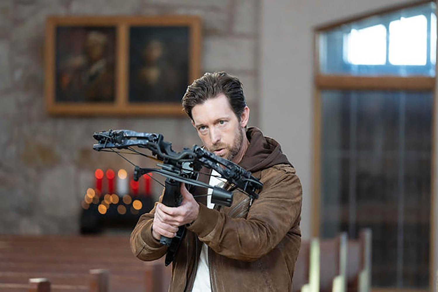 Ein Mann mit Armbrust in einer Kirche, Kerzen und Gemälde im Hintergrund.