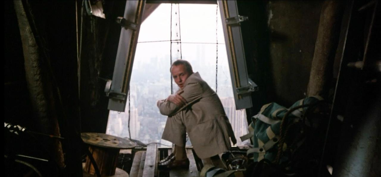 Mann in beigem Mantel sitzt vor Fenster mit Blick auf Stadt.