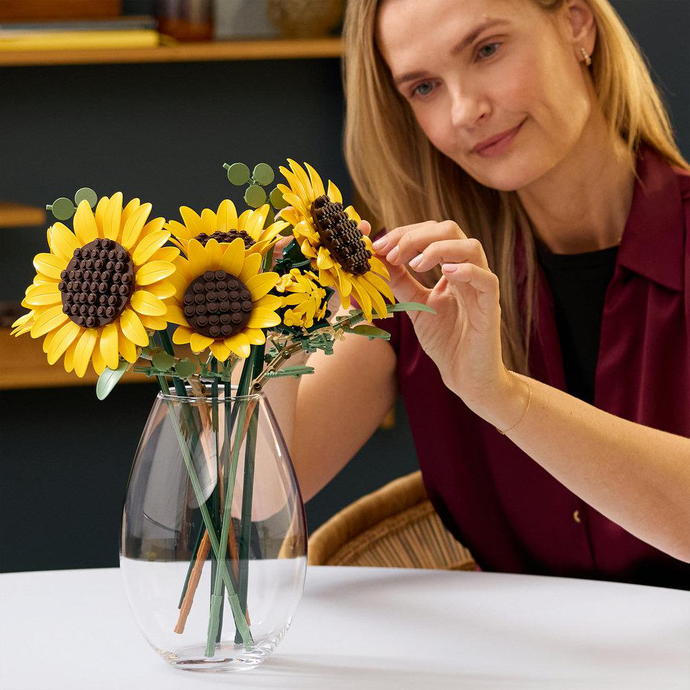 Frau arrangiert LEGO-Sonnenblumen in einer Glasvase auf einem Tisch.
