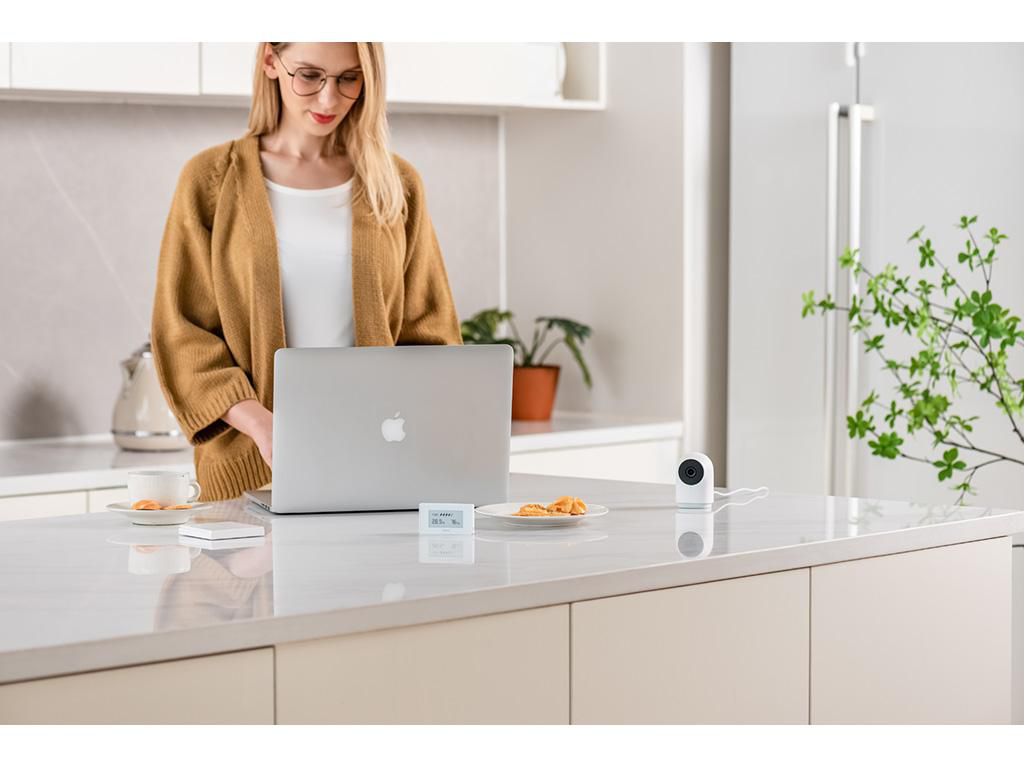 Frau im braunen Cardigan benutzt Laptop in der Küche, neben Kaffeetasse und Sicherheitskamera.