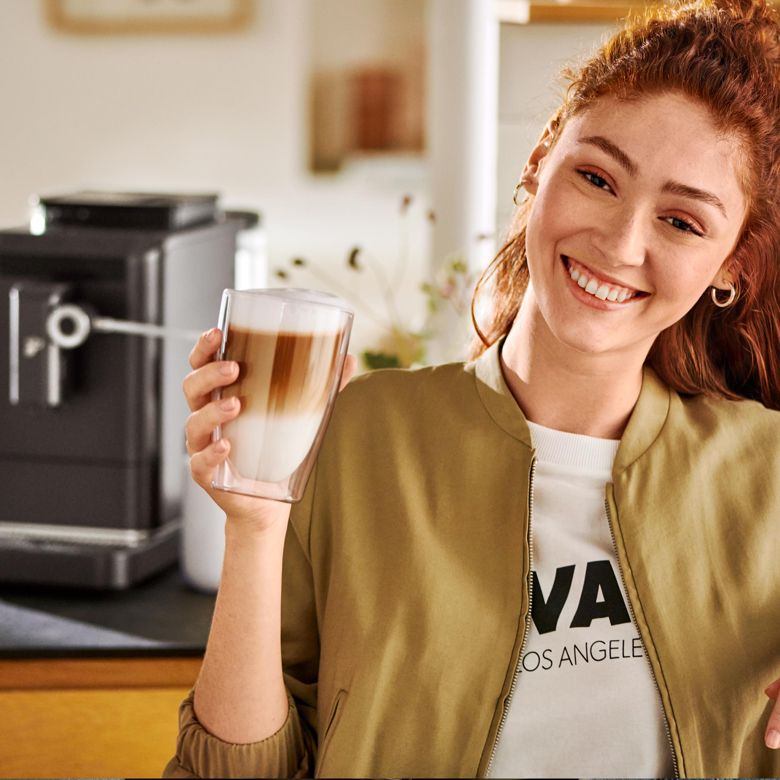 Frau mit roten Haaren hält ein Glas Latte Macchiato vor einer Kaffeemaschine.