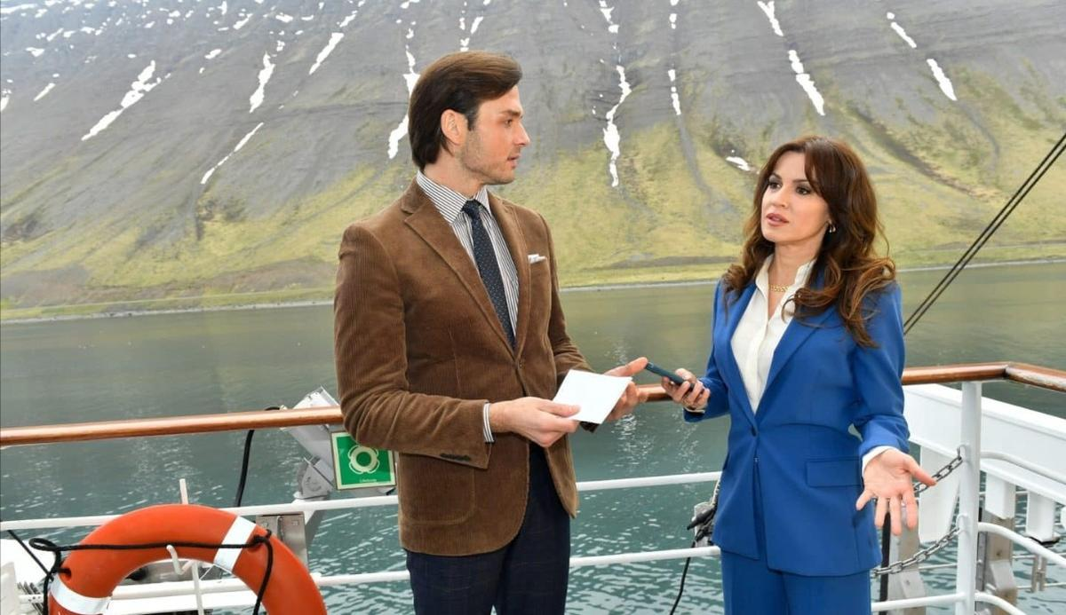 Mann in braunem Sakko und Frau in blauem Anzug auf einem Schiff vor Bergen.