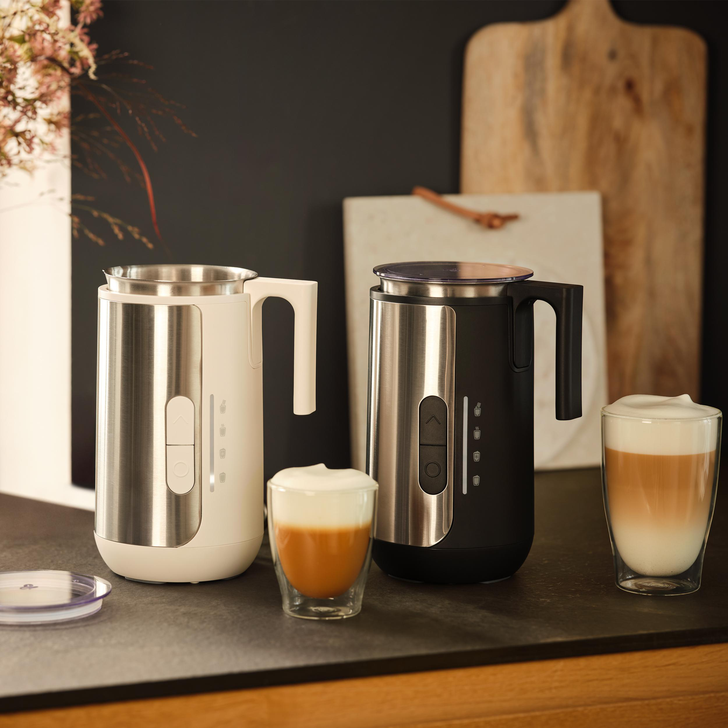 Zwei Milchaufschäumer, einer weiß, einer schwarz, mit Gläsern Kaffee mit Milchschaum.