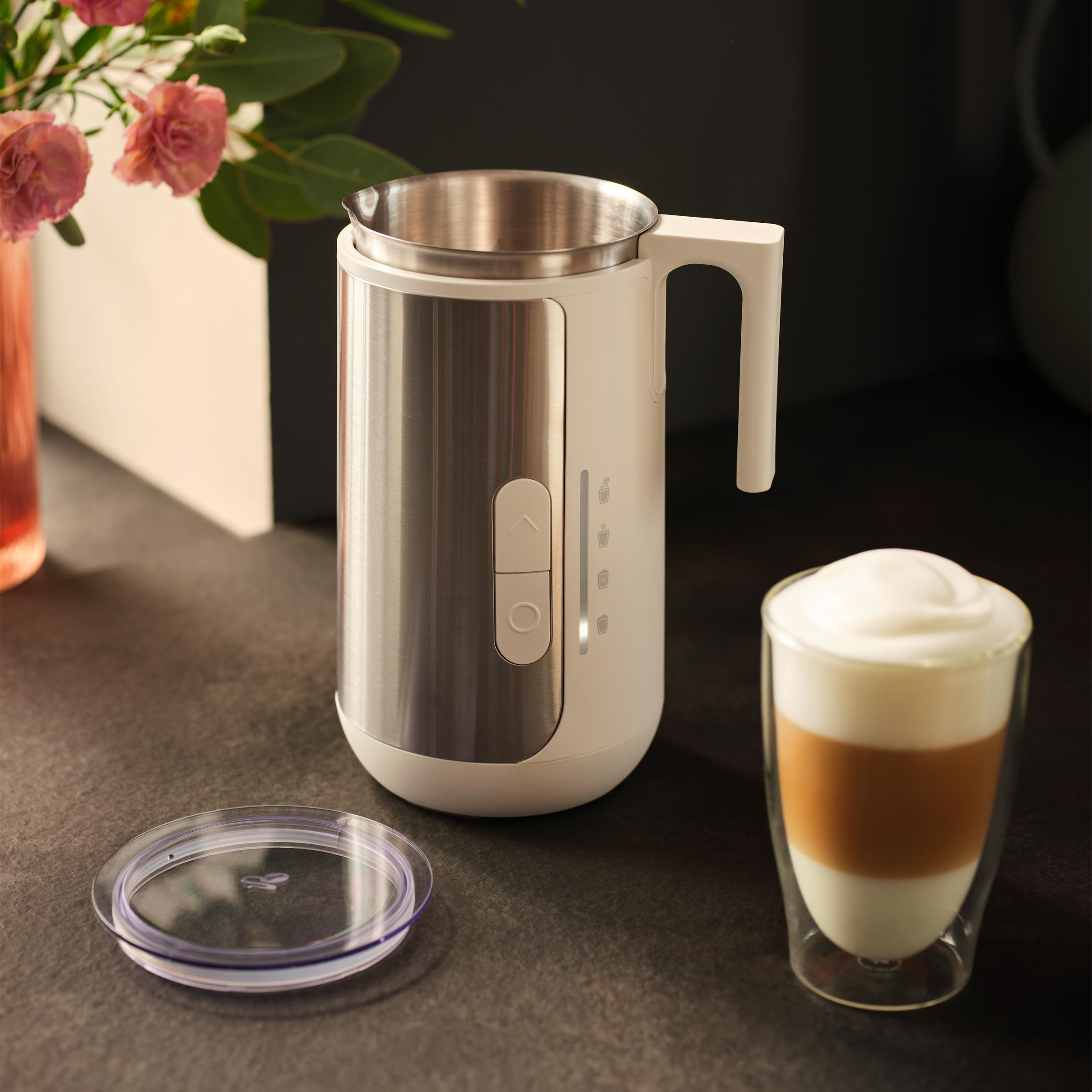 Ein Milchaufschäumer mit Edelstahlkrug, daneben ein Glas Latte Macchiato.