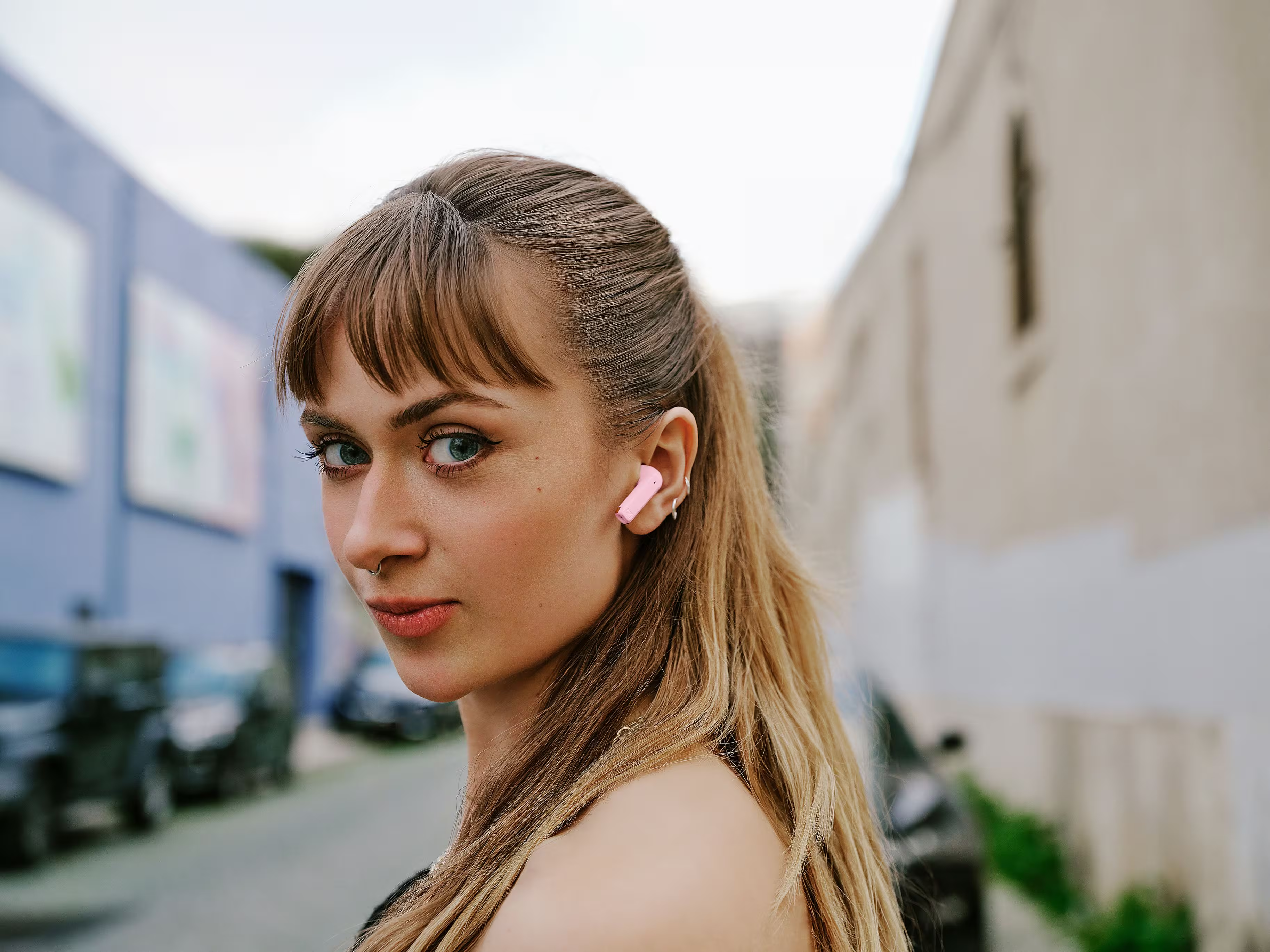 Une jeune femme aux cheveux blonds porte un écouteur rose et regarde la caméra.