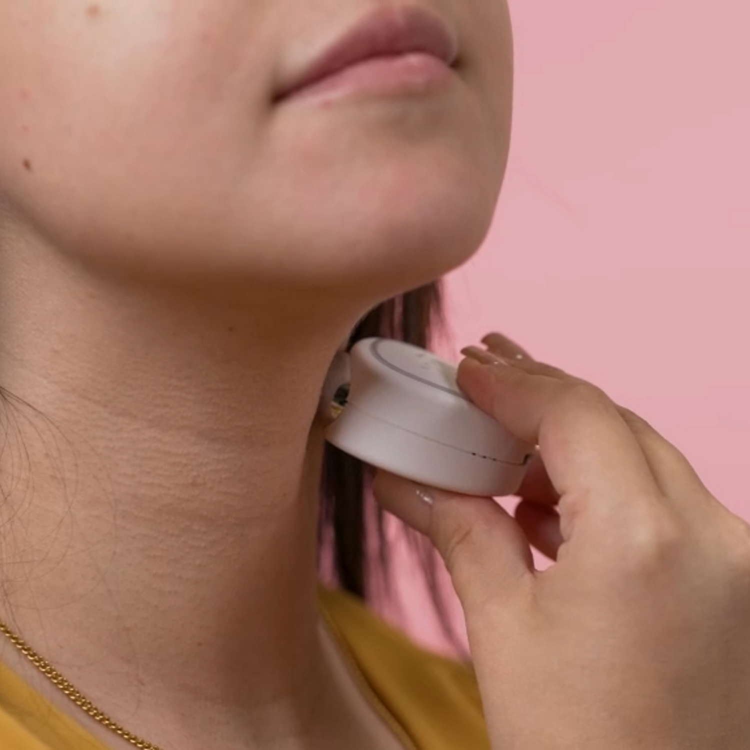 Una mano sostiene un dispositivo blanco en el cuello de una persona. Fondo rosa.