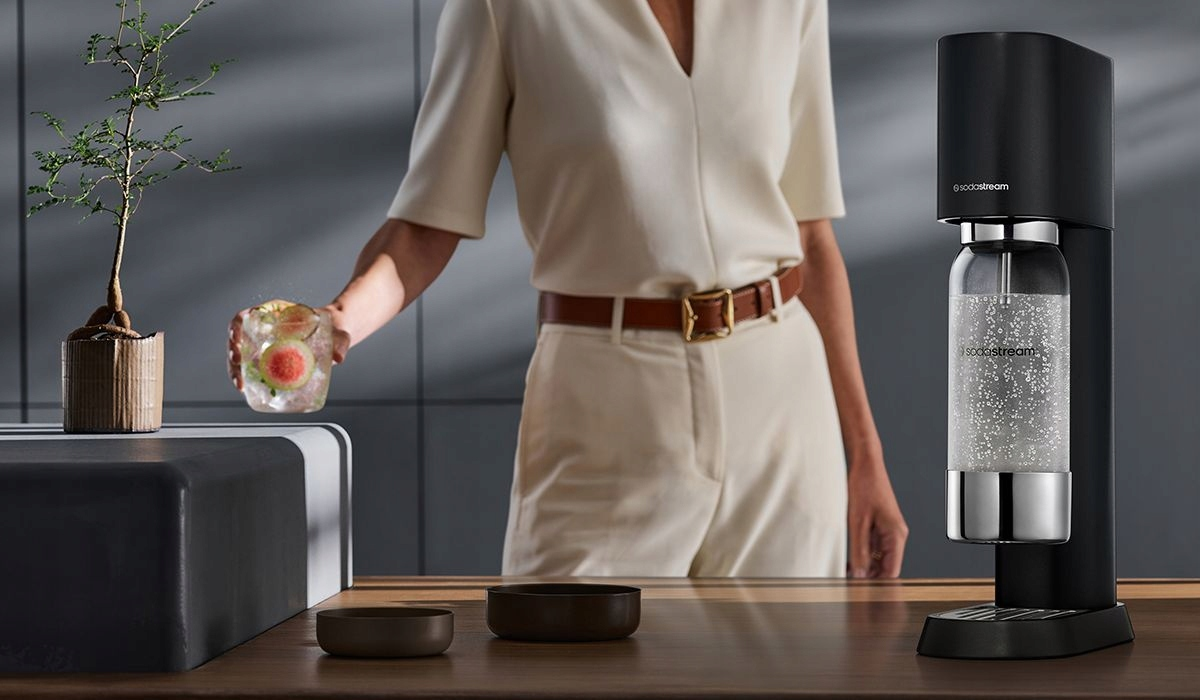 Osoba trzyma szklankę z lodem i owocami obok urządzenia SodaStream.