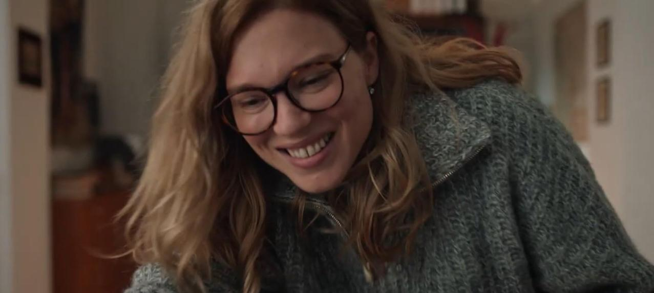 Eine Frau mit blonden Haaren und Brille lächelt in einem gestrickten Pullover.
