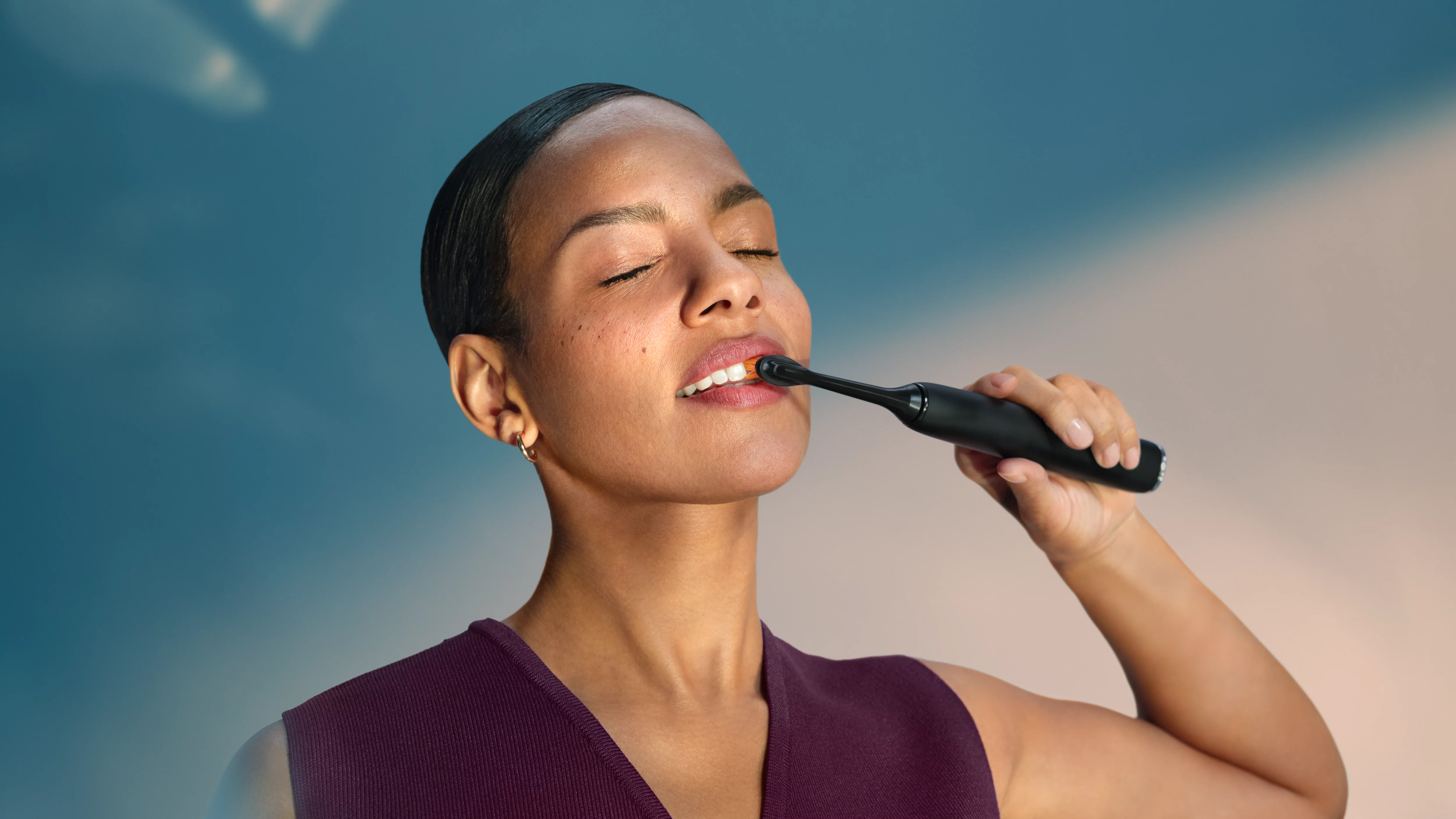 Kobieta myje zęby czarną szczoteczką elektryczną na tle niebieskiego nieba.
