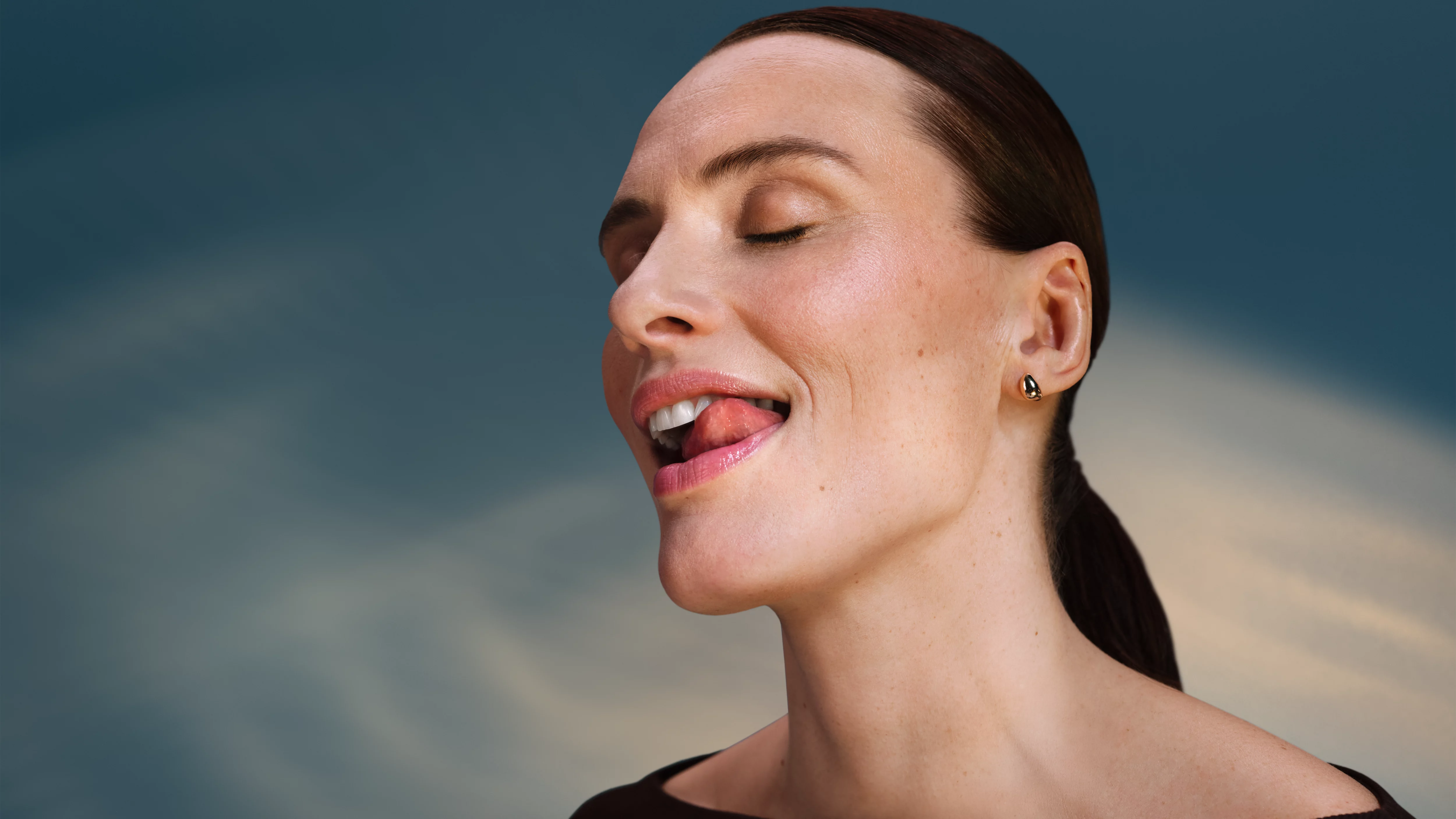 Kobieta z zamkniętymi oczami liże usta, złoty kolczyk.