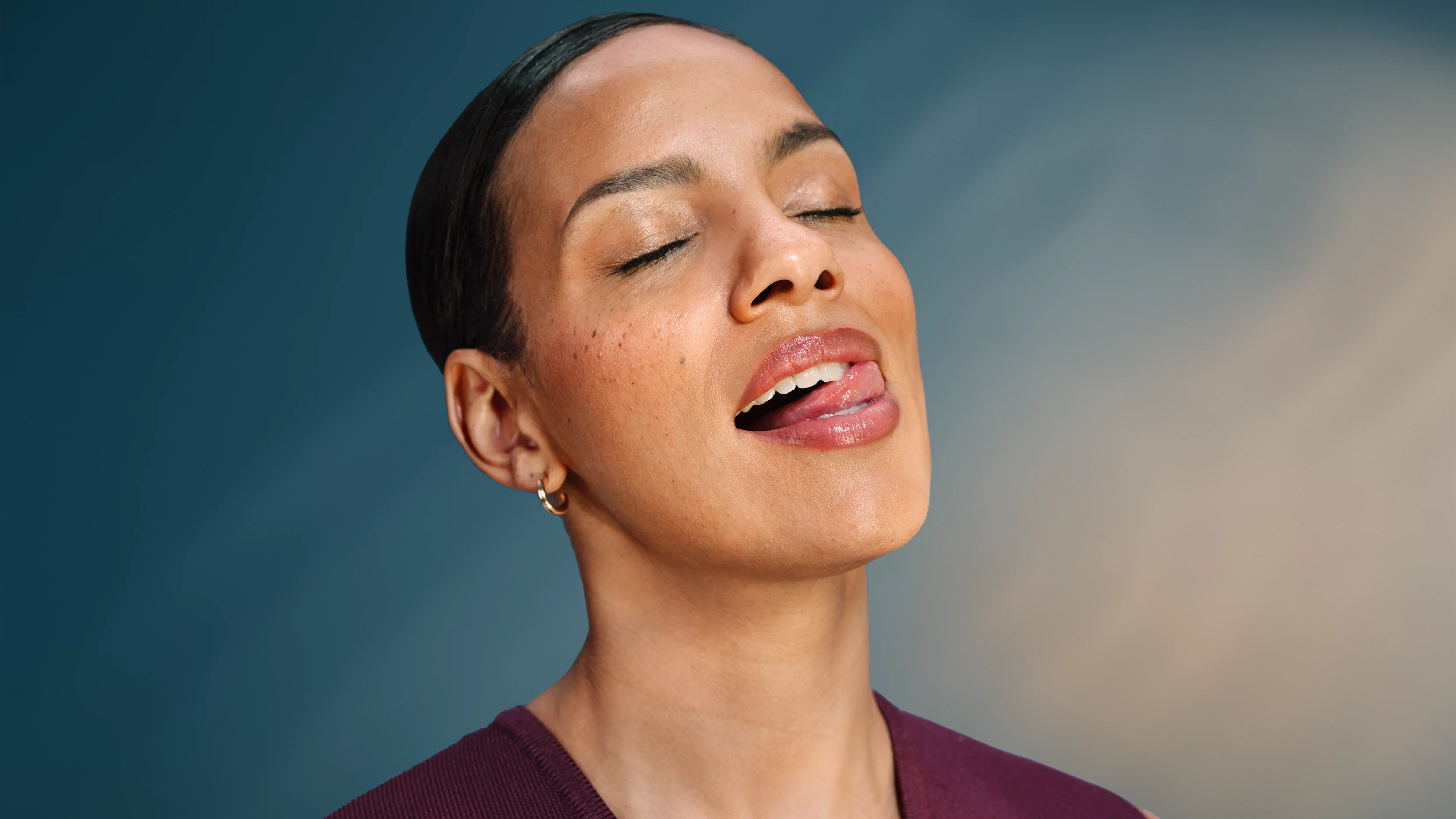 Kobieta z zamkniętymi oczami, wystawionym językiem, błyszczącymi ustami, niebieskie tło.