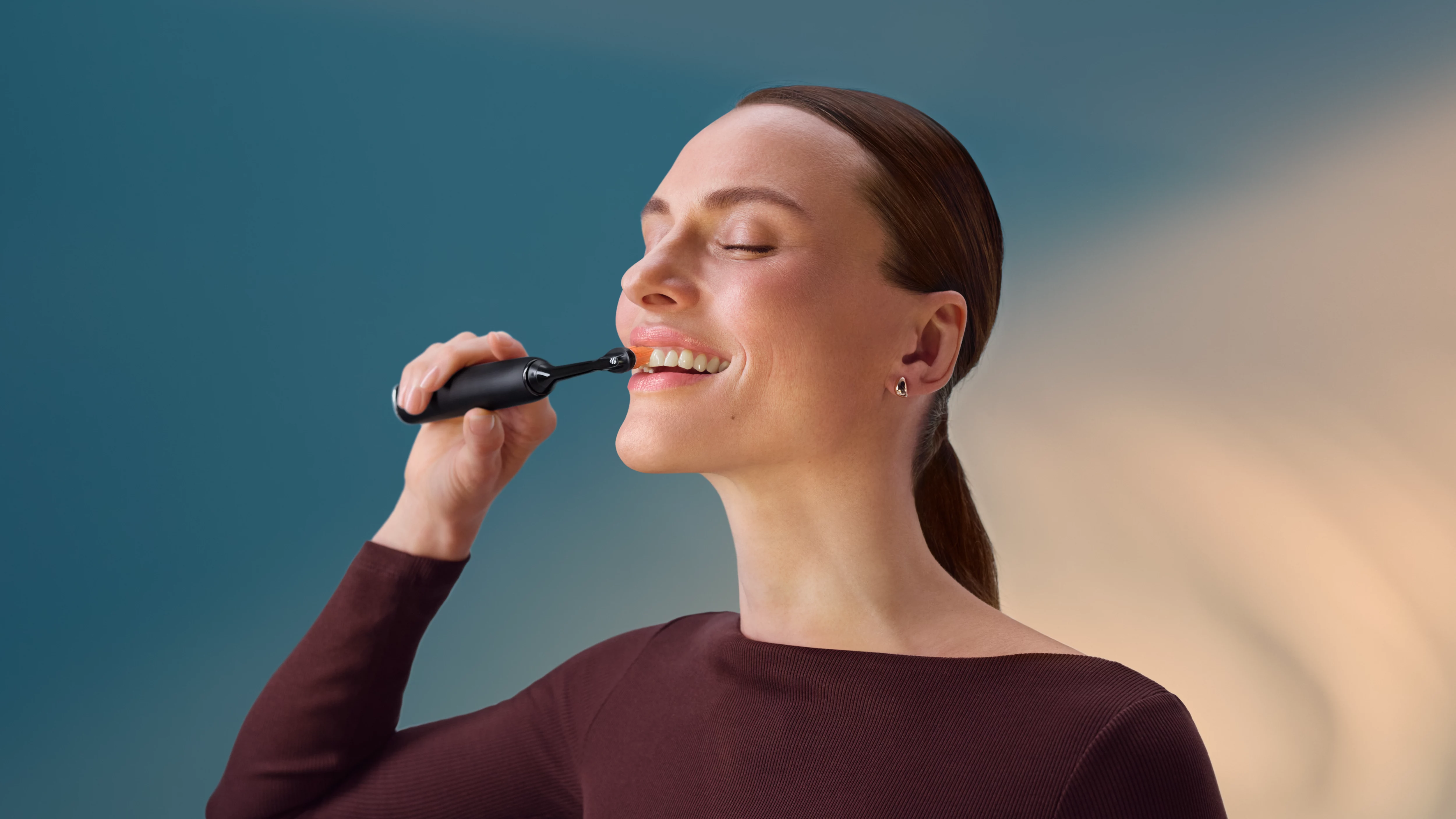 Kobieta myje zęby czarną szczoteczką elektryczną na tle niebiesko-beżowym.
