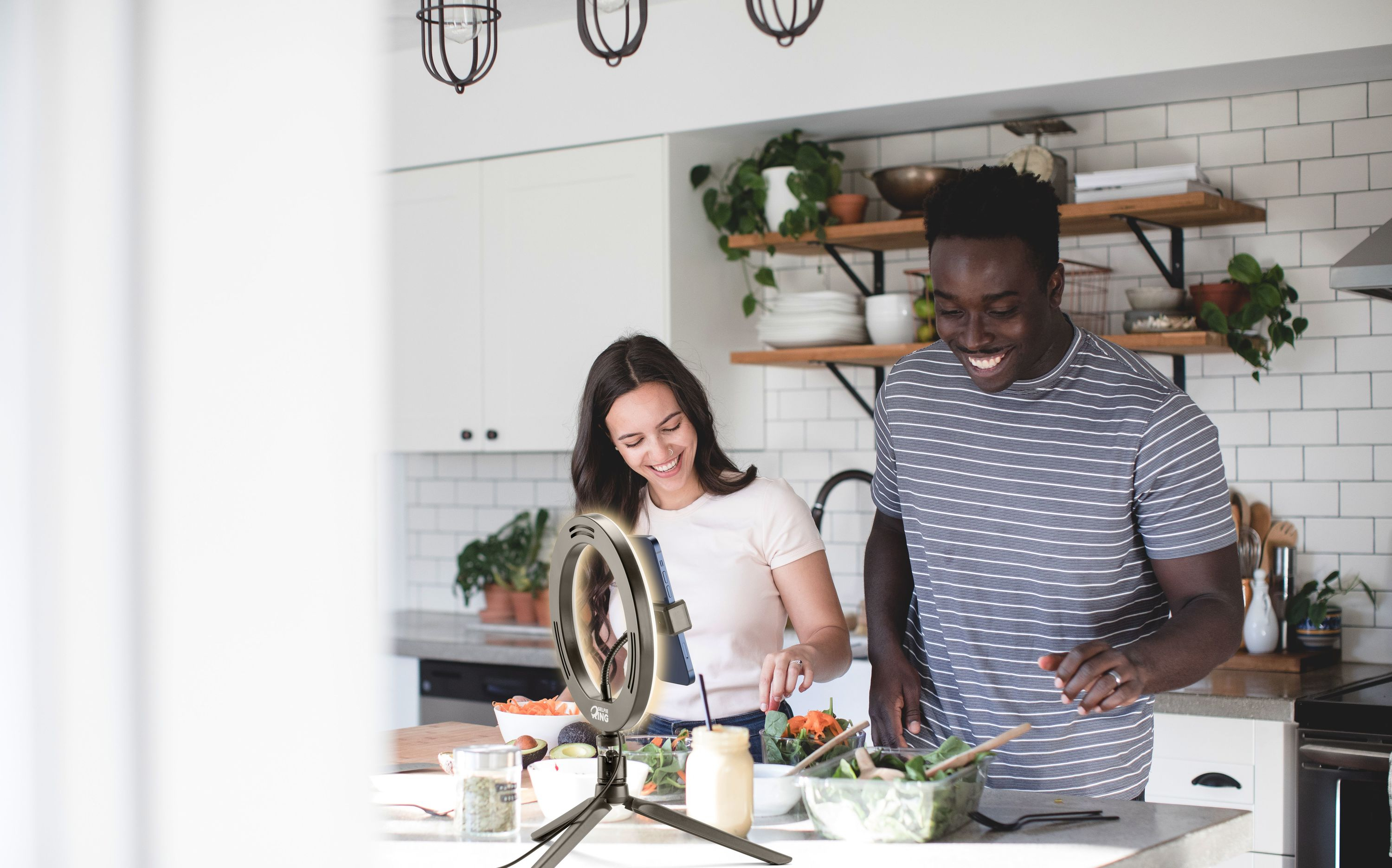 Una pareja prepara ensaladas en una cocina luminosa mientras filma una cámara con aro de luz.