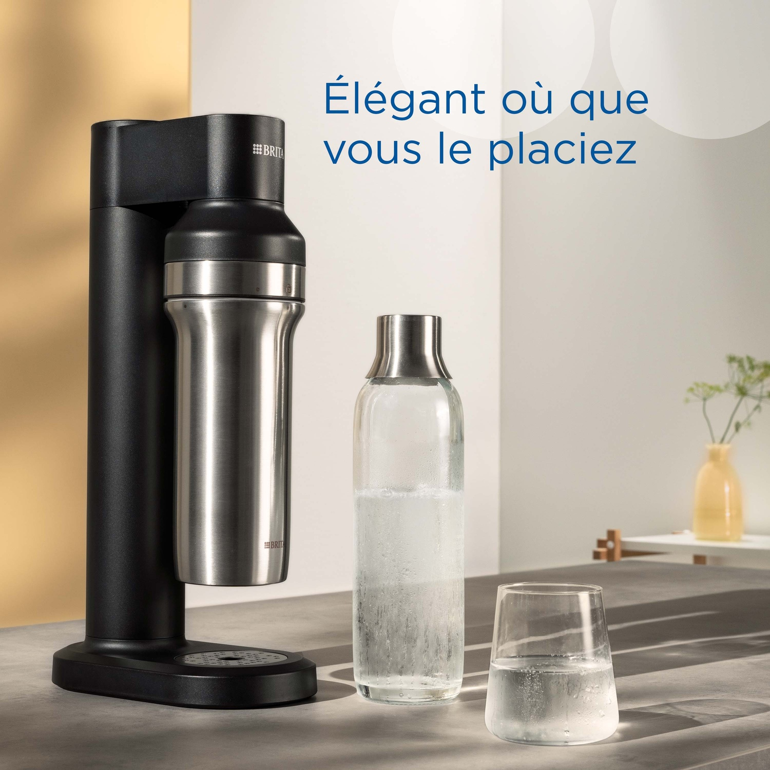 Un appareil à gazéifier l'eau noir avec une bouteille argentée, à côté une bouteille en verre et un verre d'eau.