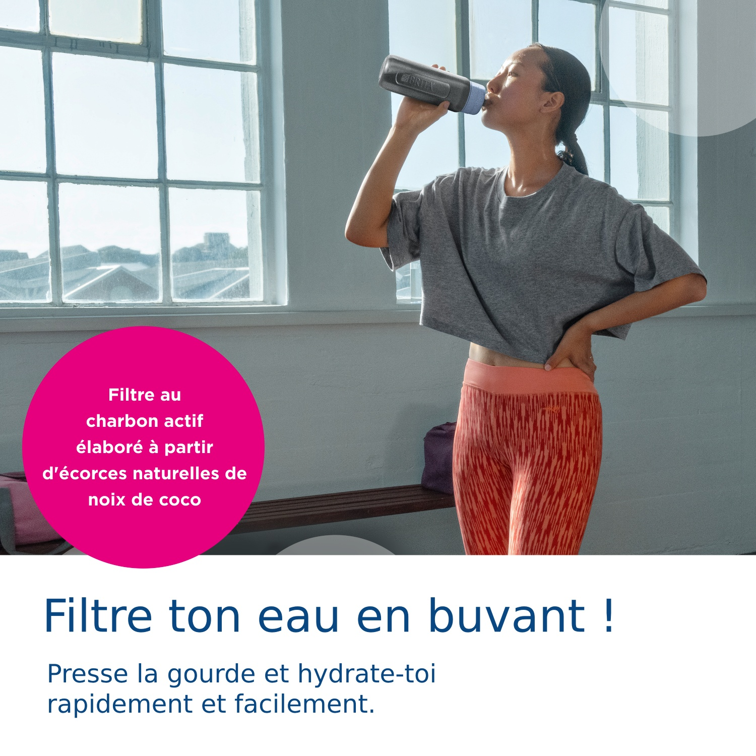 A woman drinks from a gray water bottle in a bright room with large windows.