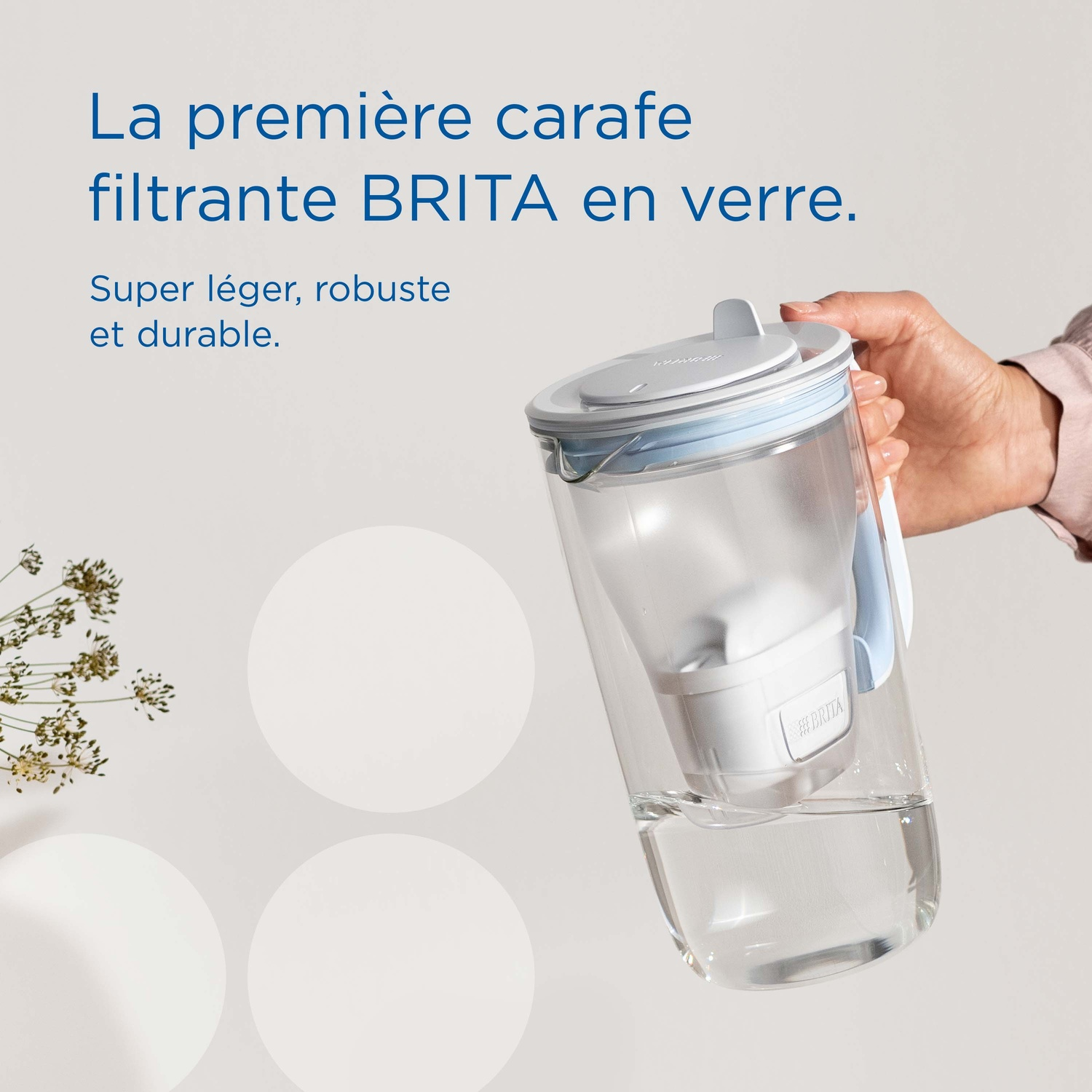 A hand holds a clear glass BRITA pitcher with a blue lid and filter.