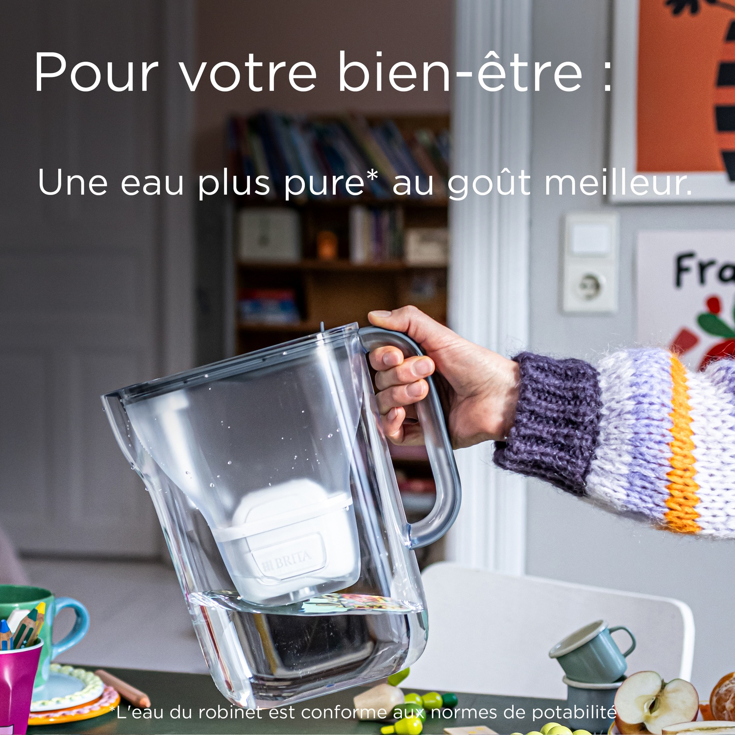 Une main tient une carafe Brita avec de l'eau filtrée. Tasses d'enfants en arrière-plan.